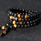 108 bead obsidian tiger eye meditation bracelet coiled 3 layers on dark surface showing engraved cylinder bead and gold accent