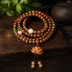 TAIJIAURA consecrated agarwood mala 108 beads laid flat in circle on dark wooden table with jade green orange accent beads carved lotus pendant purple clay teapot green leaves spirit gathering deep meditation focus