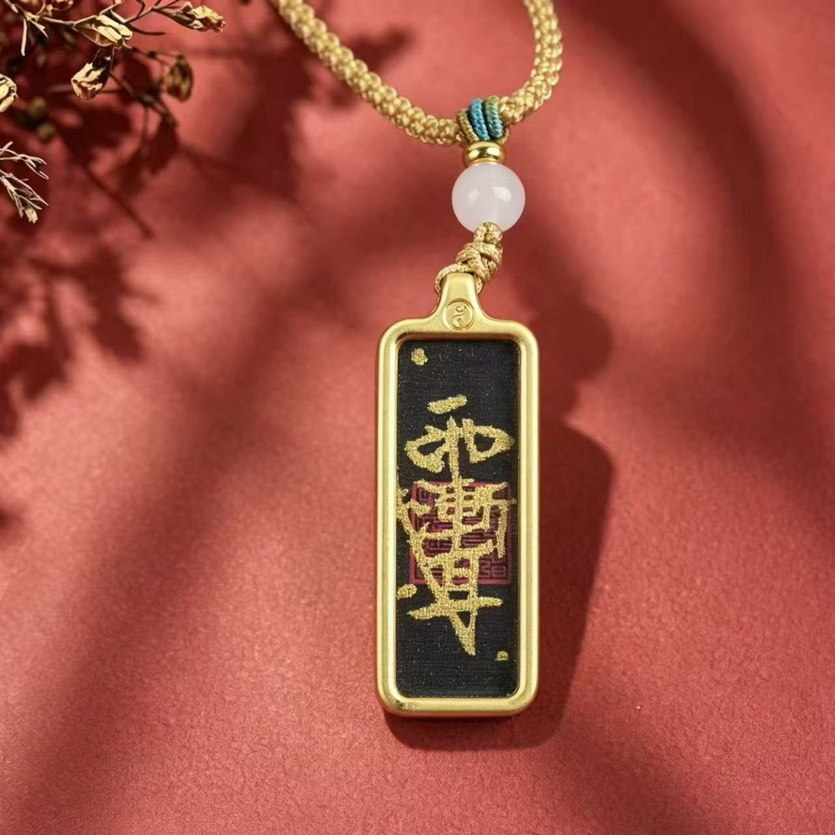 Front view of a black Taoist talisman pendant with a gold-colored frame, woven cord and pale round bead on a red background.