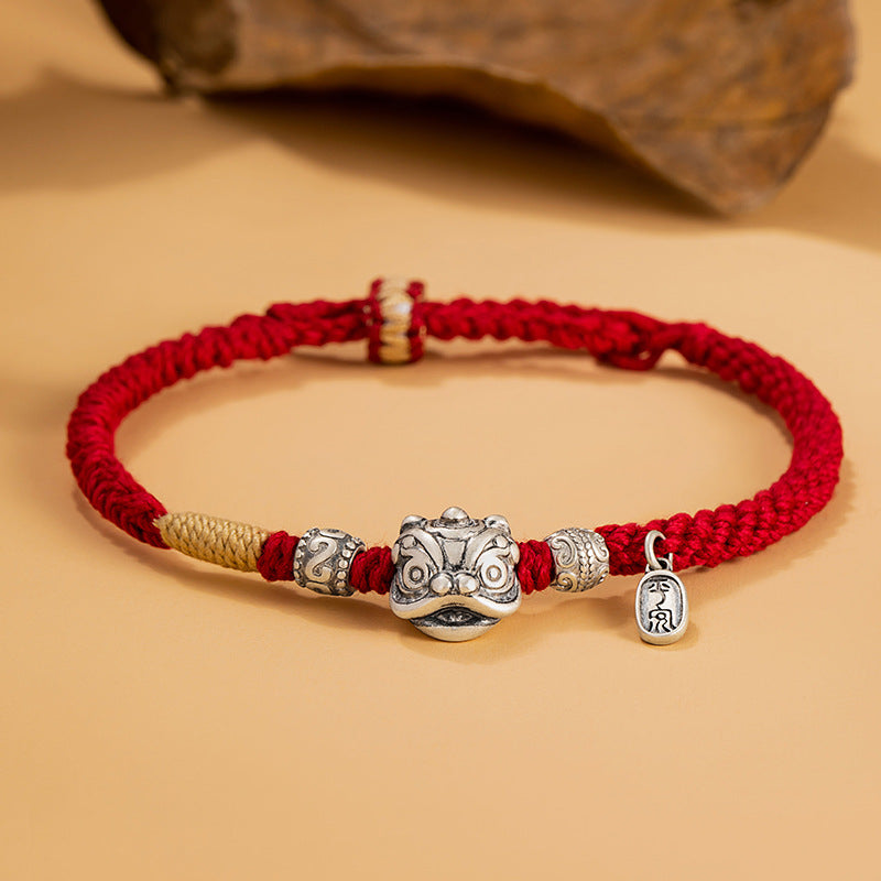 Red braided cord bracelet with silver lion head charm and rune beads on warm stone background showing Taoist KaiGuang love protection couple jewelry detail