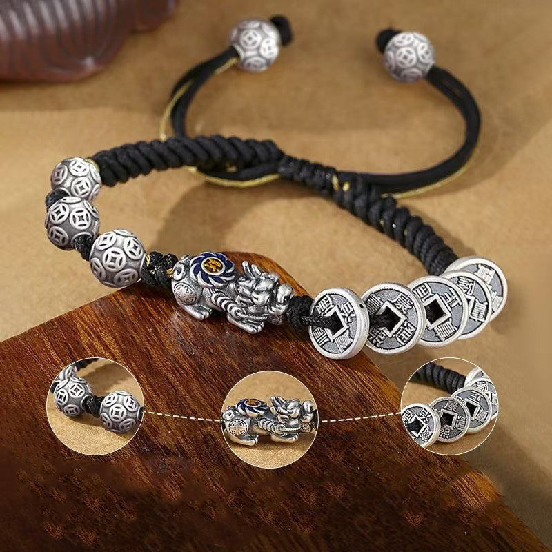 Silver Pixiu with blue enamel eyes and five ancient coin charms on black braided wealth bracelet displayed on warm wood surface with detail closeups