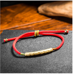 Handwoven red rope evil ward bracelet with engraved gold mantra tube on dark stone surface