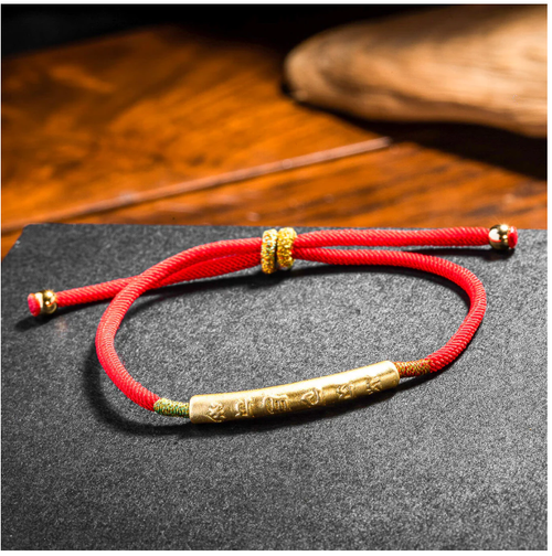 Handwoven red rope evil ward bracelet with engraved gold mantra tube on dark stone surface