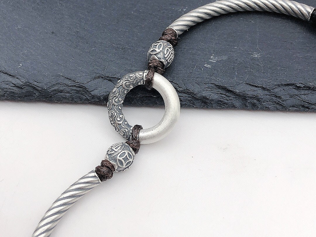 Close-up of white jade peace buckle ring carved silver cloud pattern surround engraved silver beads on silver twisted rope brown braided cord slate background Taoist KaiGuang protection evil warding amulet detail
