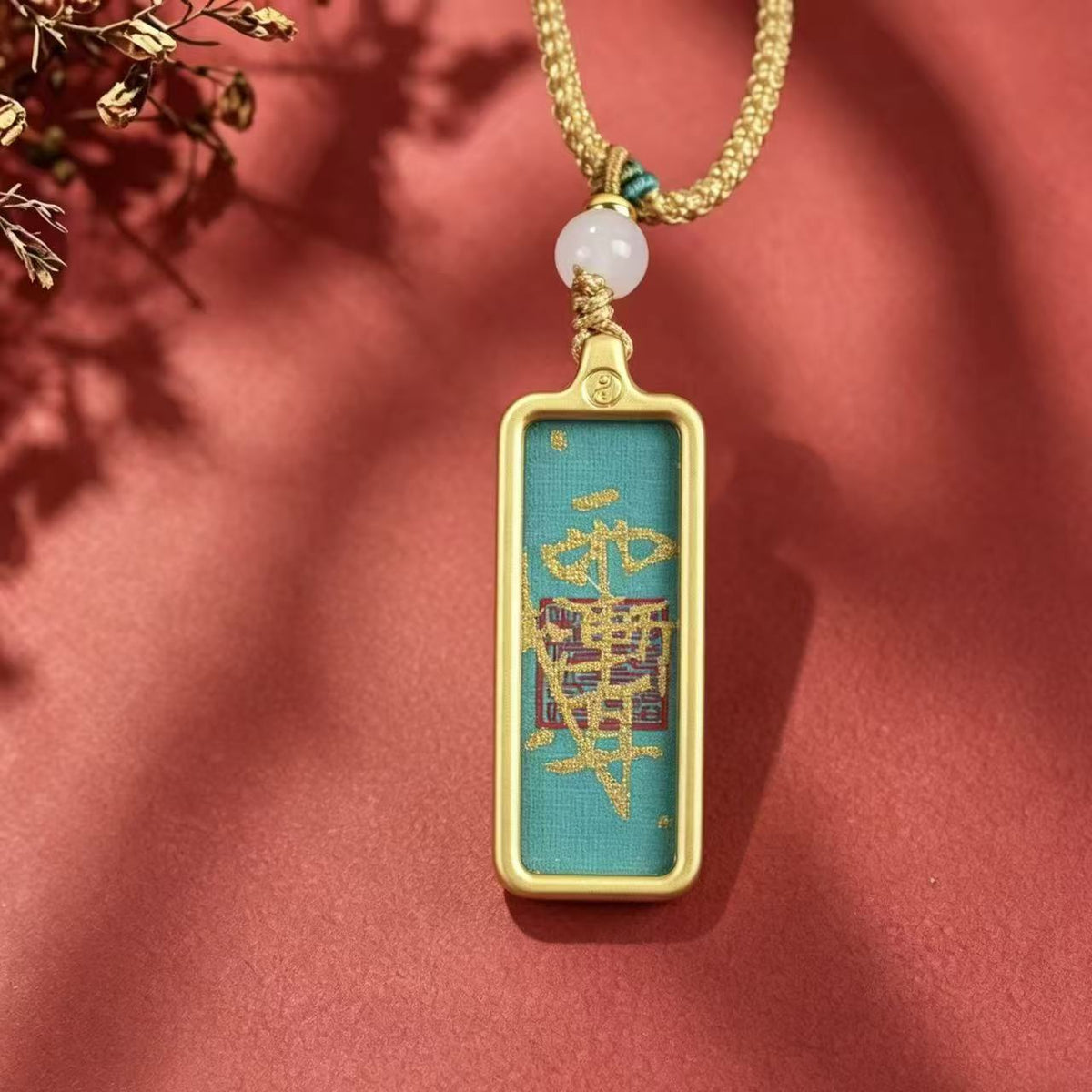 Front view of a teal Taoist talisman pendant with gold-colored markings and frame on a red background.